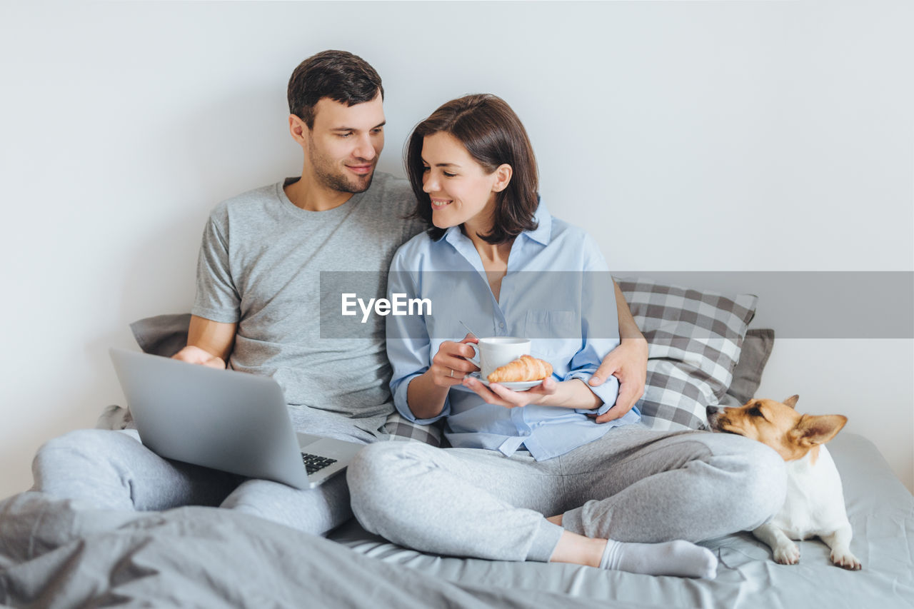 Couple watching video over laptop while sitting with dog on bed against wall at home