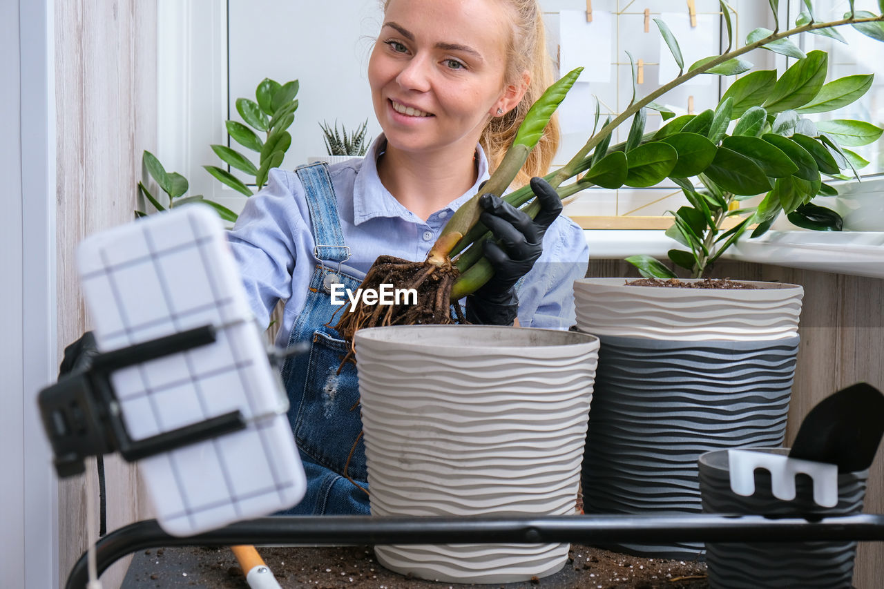 Gardener woman blogger using phone while transplants indoor plants and use a shovel on table. 