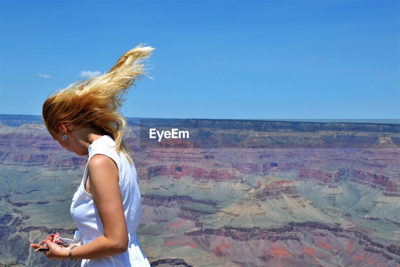 Side view of woman with tousled blond hair standing at grand canyon national park against blue sky