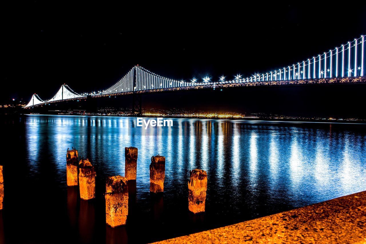 ILLUMINATED BRIDGE OVER RIVER AGAINST SKY AT NIGHT