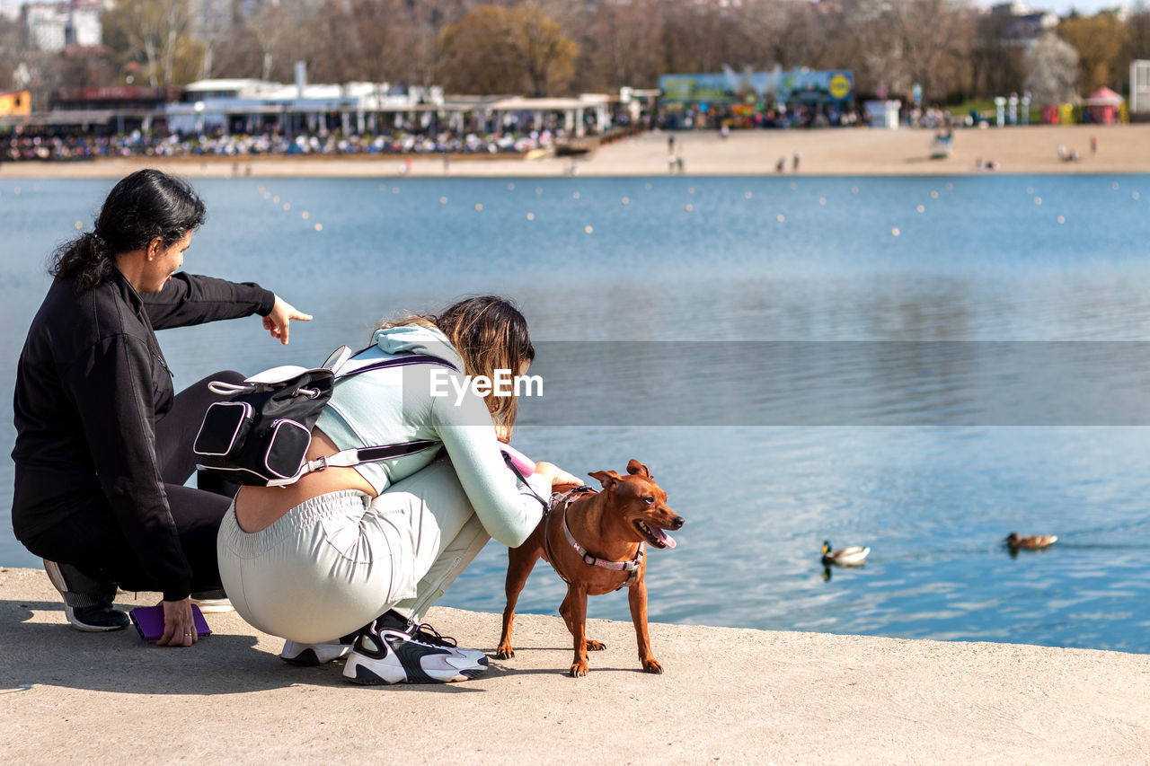 Young girl and her mother on relaxing vacation by the lake with their cute miniature pinscher.