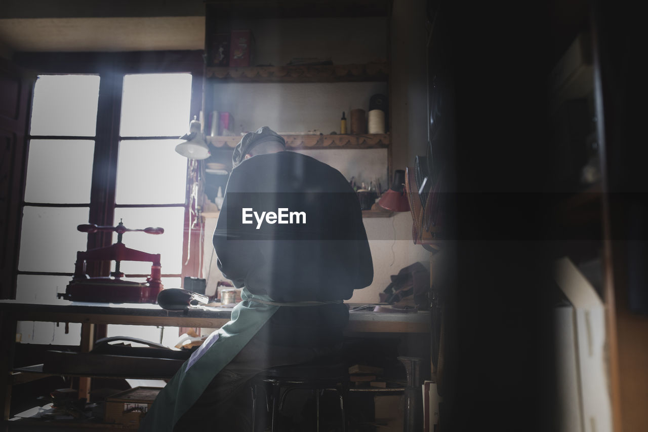 Back view of unrecognizable craftsman sitting working in dark workshop