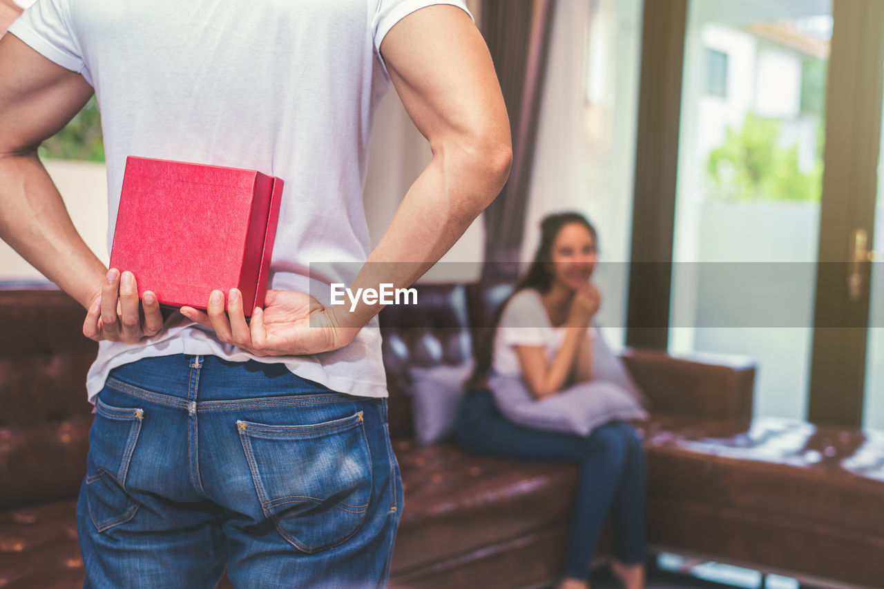 Midsection of boyfriend hiding gift while girlfriend sitting on sofa at home