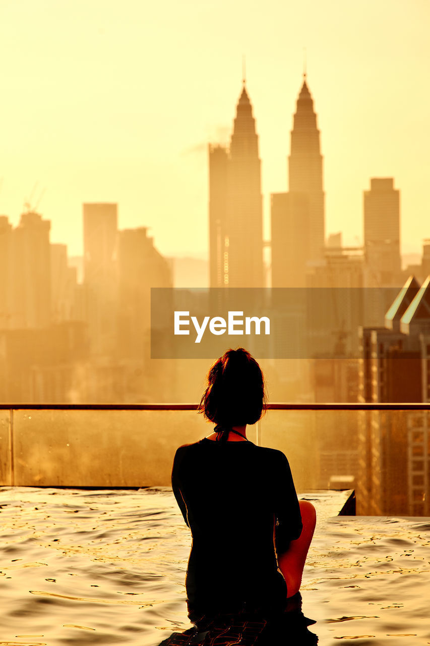 Rear view of woman sitting swimming in infinity pool during sunset