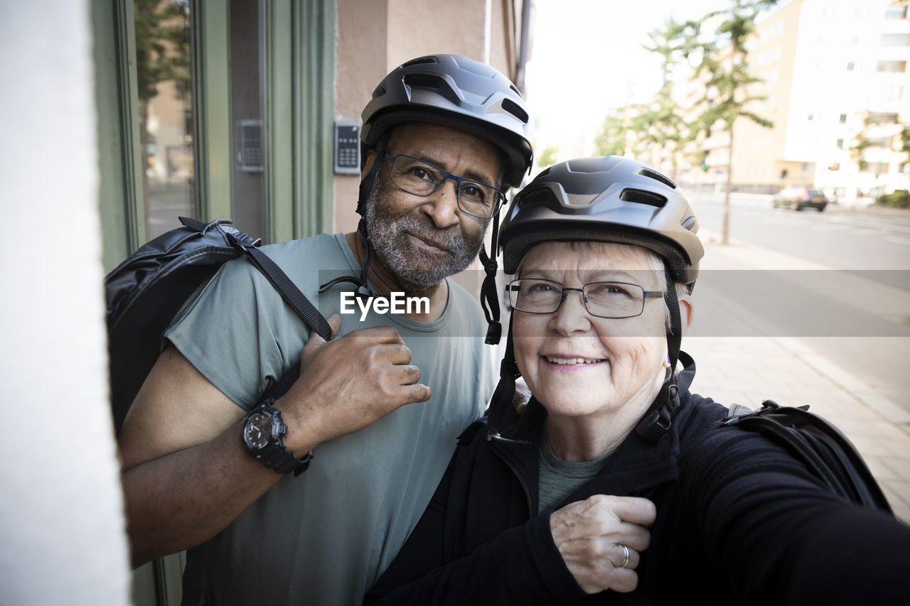 Smiling senior couple taking selfie with cycling helmet outside house