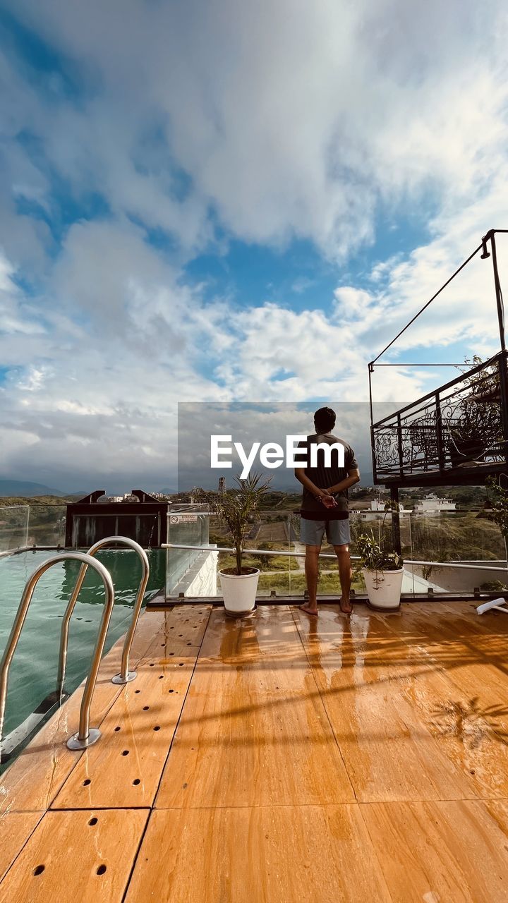 sky, cloud, one person, water, vacation, full length, nature, sea, men, adult, standing, railing, wood, architecture, rear view, day, leisure activity, lifestyles, outdoors, walkway, casual clothing, trip, holiday, travel, boardwalk, ocean, sunlight, deck, sports, young adult, relaxation, built structure