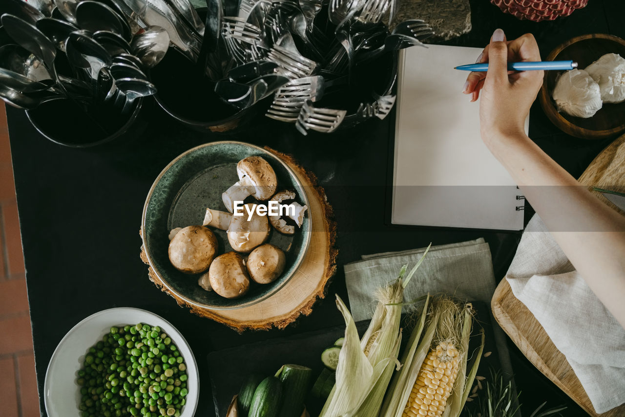 Hand of female chef writing recipe in diary by vegetables and cutlery on counter