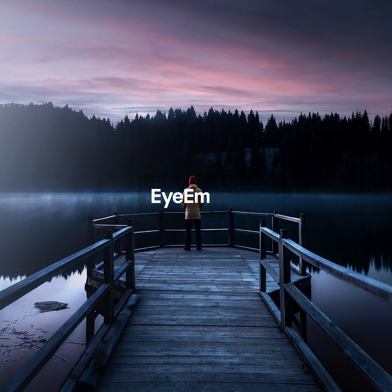 Rear view of man standing on pier over lake against sky during sunset