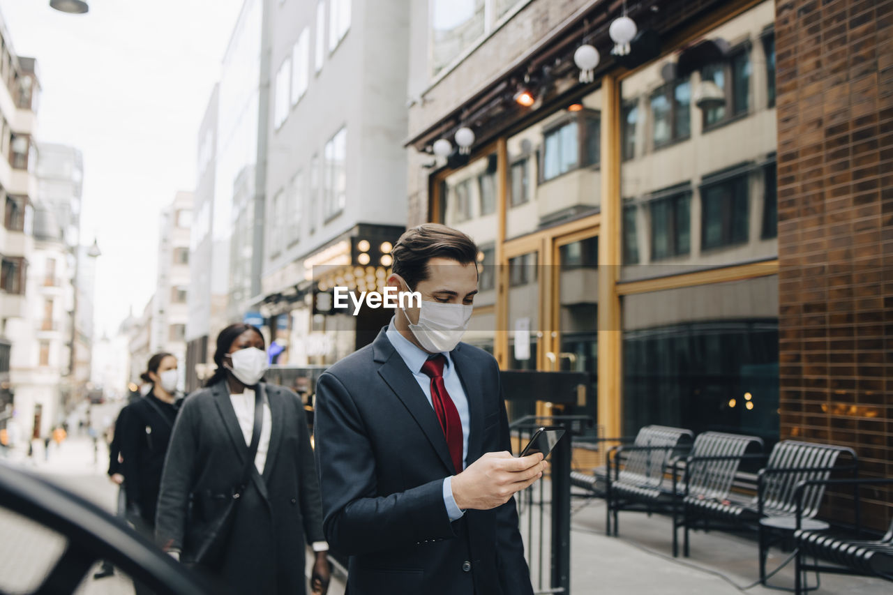 Businessman using smart phone while female colleagues social distancing in city during pandemic