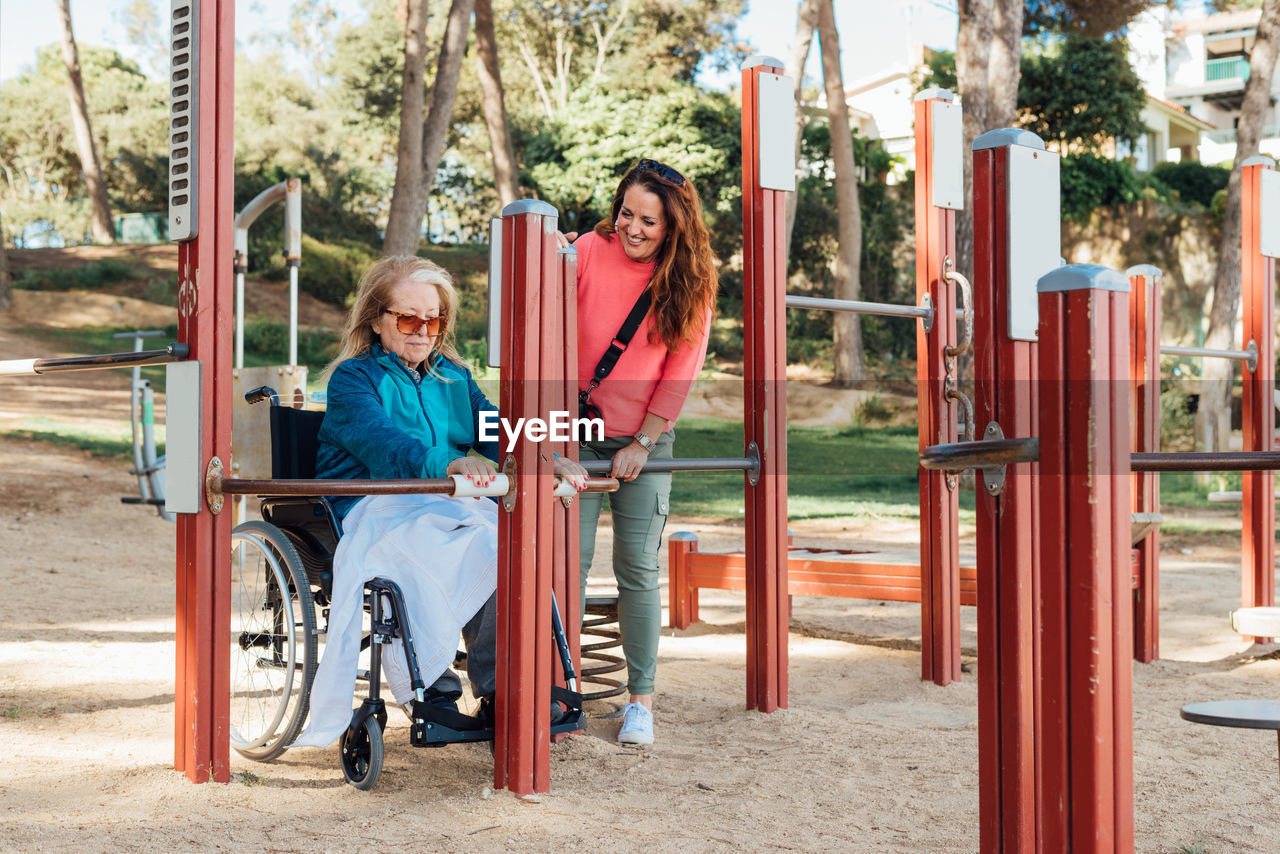 Content adult woman helping senior mother in wheelchair doing exercises on sports ground during rehabilitation session