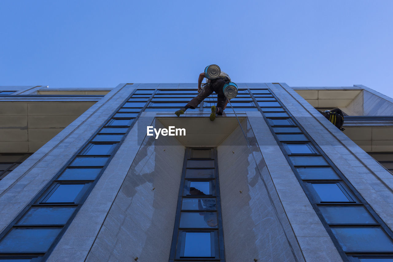 Low angle view of window washers working on building against clear blue sky