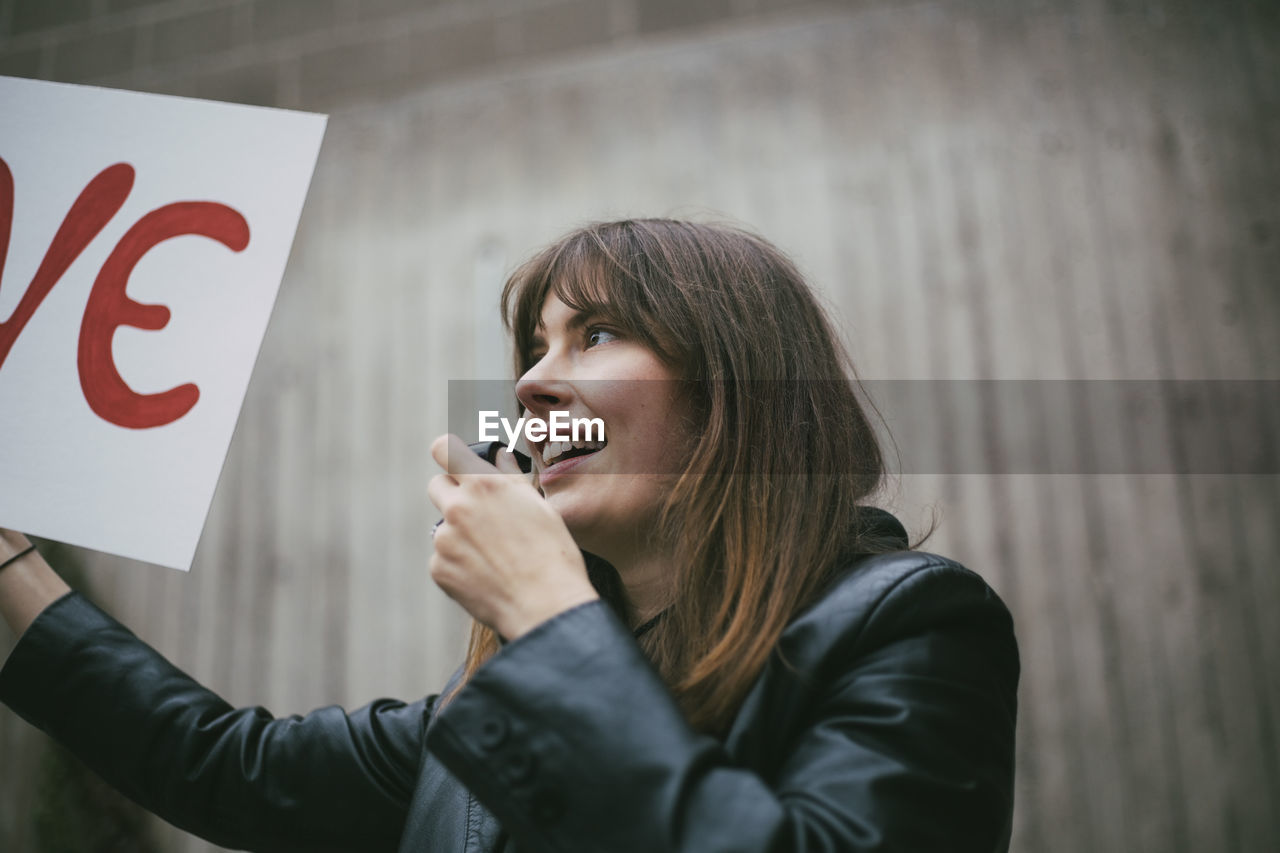 Smiling female activist with whistle holding poster against wall