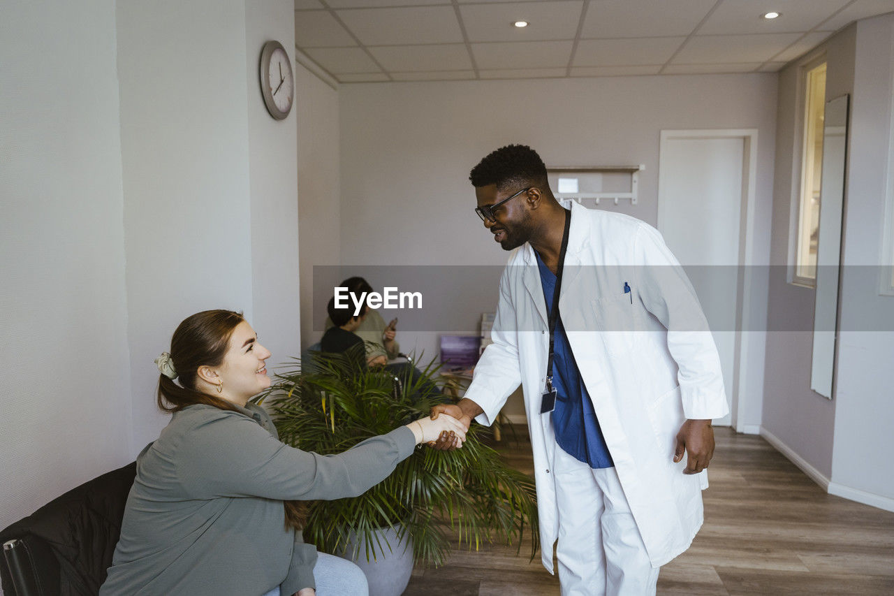 Smiling male doctor greeting female patient sitting in waiting room at clinic