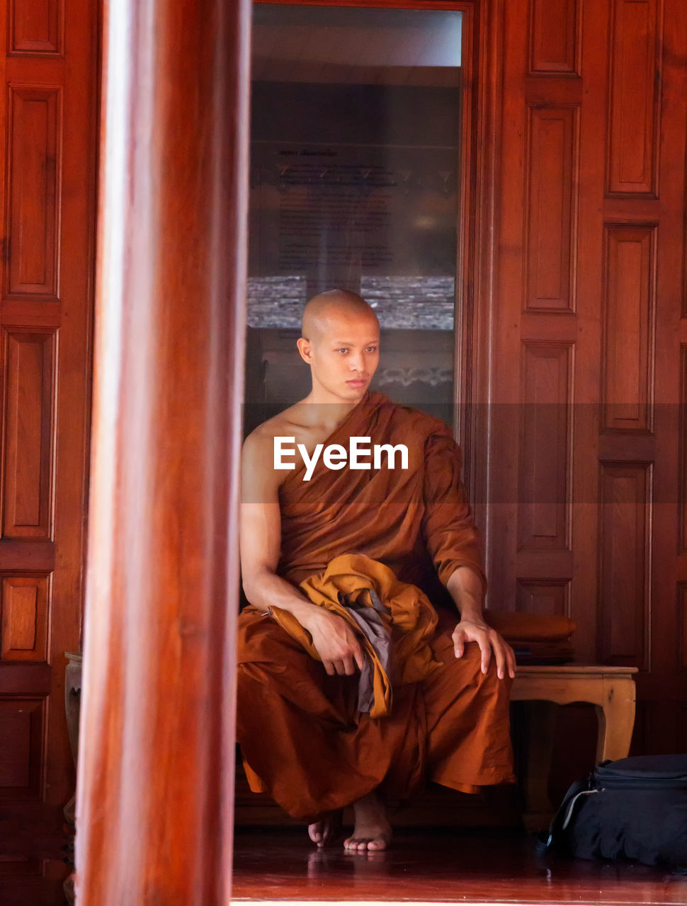 Full length of monk wearing traditional clothing while sitting against building