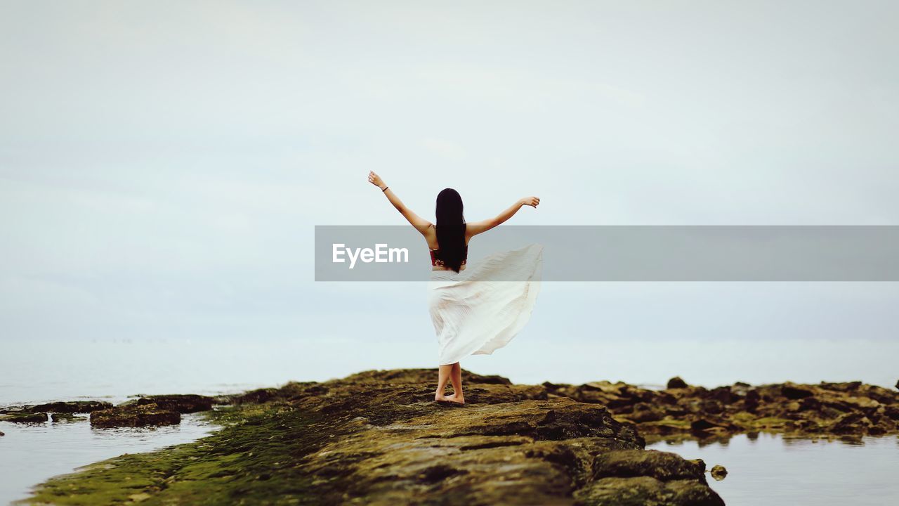 Rear view of woman standing on rock by sea against sky