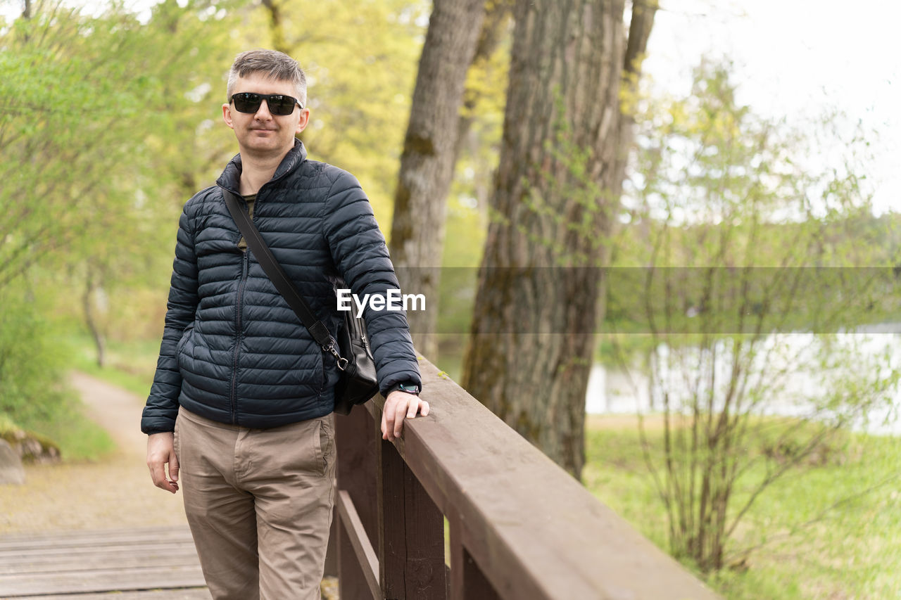 Middle-aged man in sunglasses and thin down jacket on wooden footbridge near lake in early spring