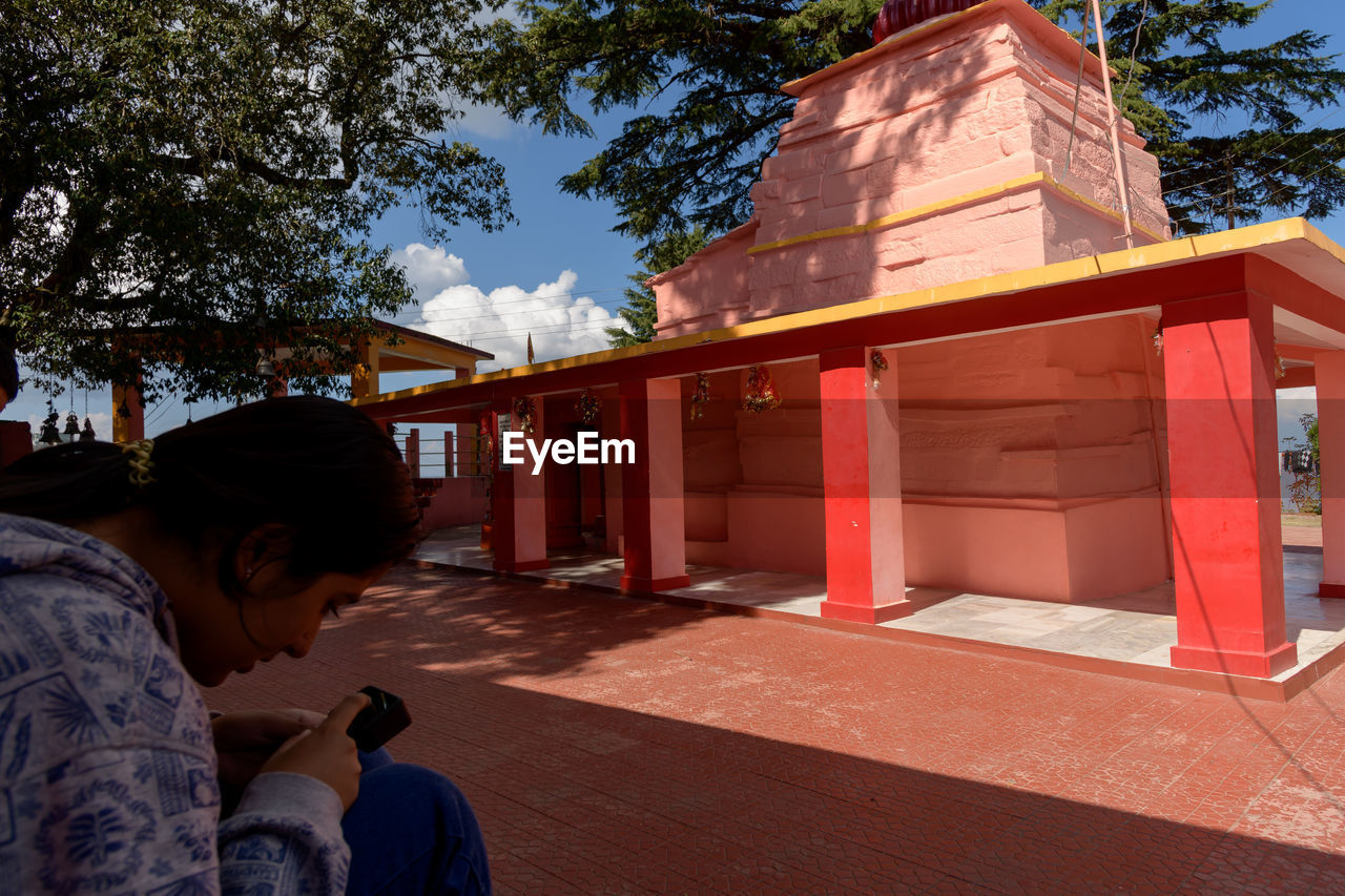 architecture, tree, built structure, nature, building exterior, plant, temple, adult, building, religion, travel destinations, one person, belief, clothing, travel, person, house, spirituality, temple - building, men, outdoors, sky, sunlight, lifestyles, city, place of worship, day, tourism, sitting