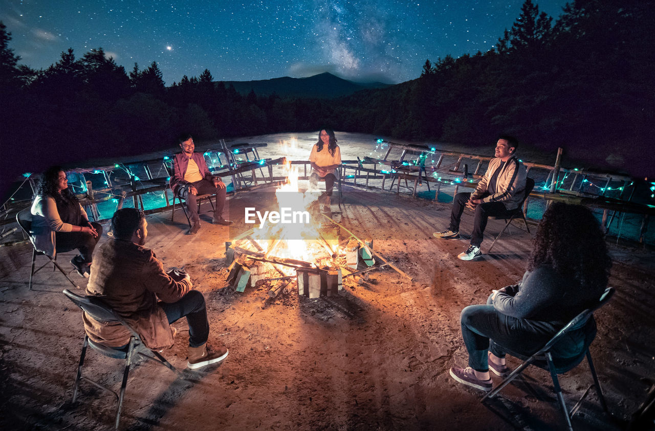 PEOPLE SITTING BY BONFIRE ON MOUNTAIN AGAINST SKY