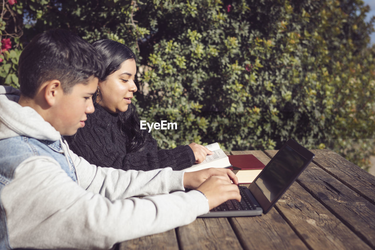 Two young latino students, outdoors, using laptops, sitting at a park