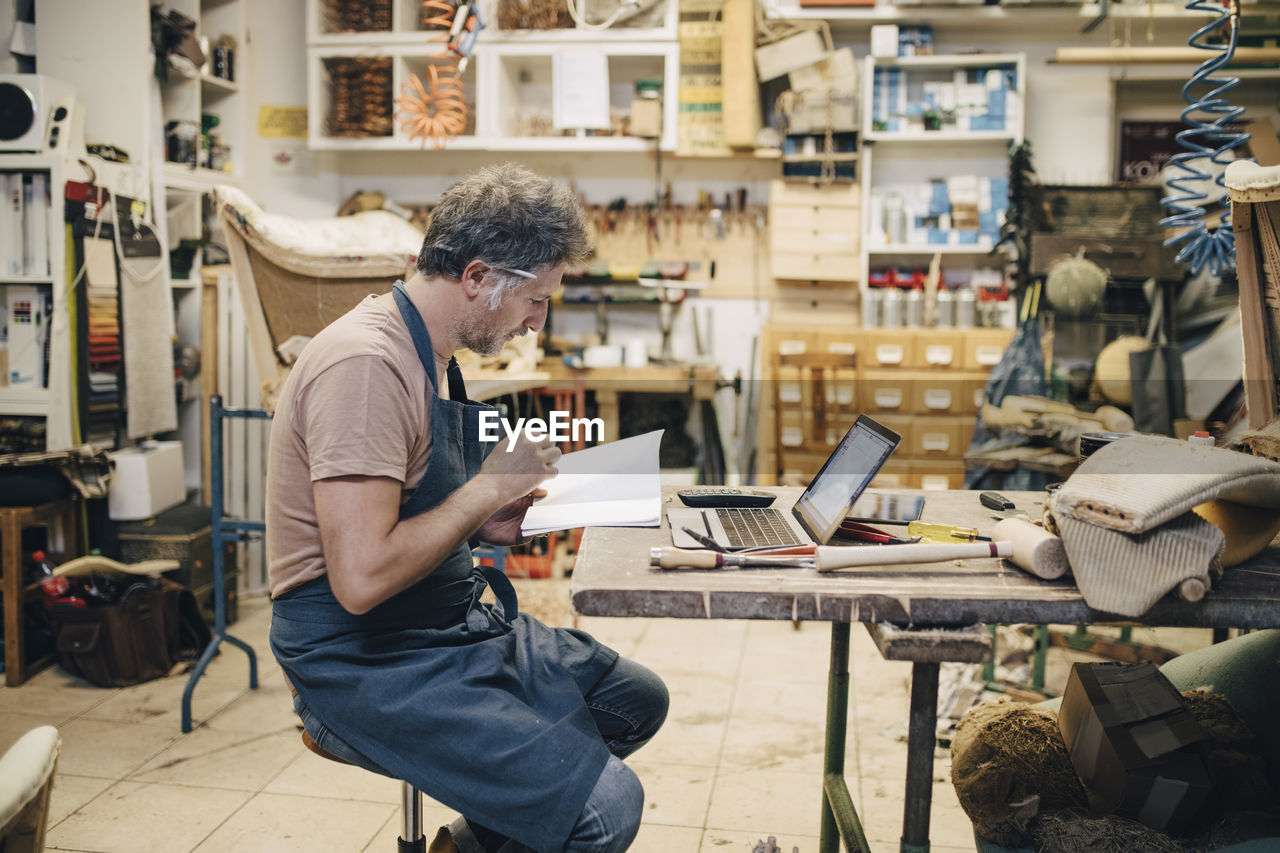 Side view of male upholstery worker reading book while sitting at workbench in workshop