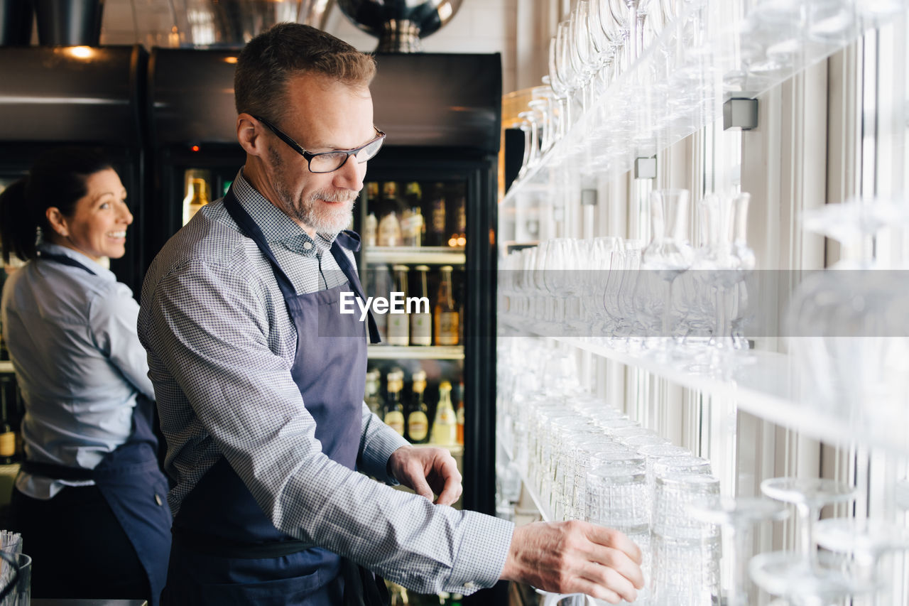 Mature man arranging wineglasses while waitress in background at restaurant