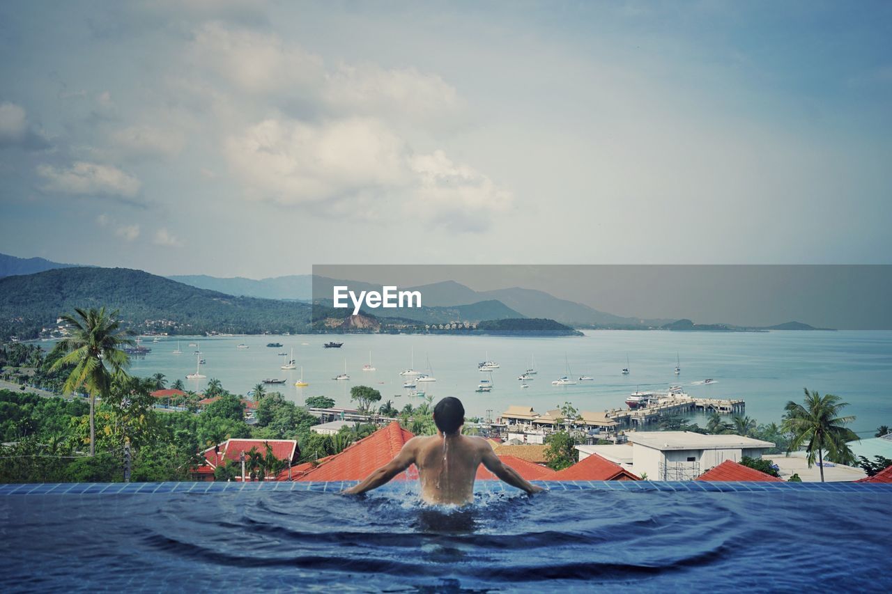 Rear view of shirtless man in infinity pool looking at sea against sky