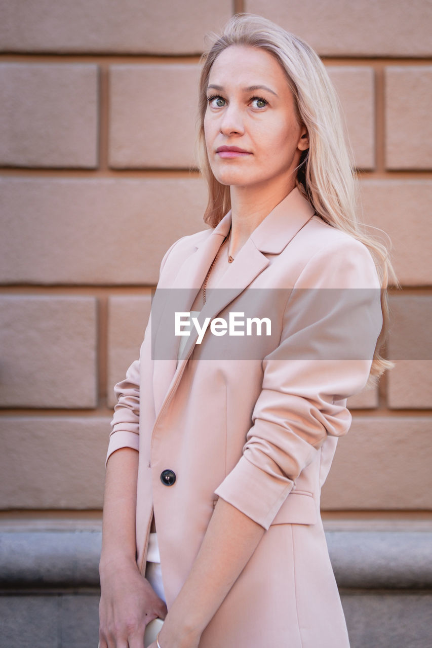 Portrait of a successful business woman with long hair, blonde. in an expensive suit, brick wall