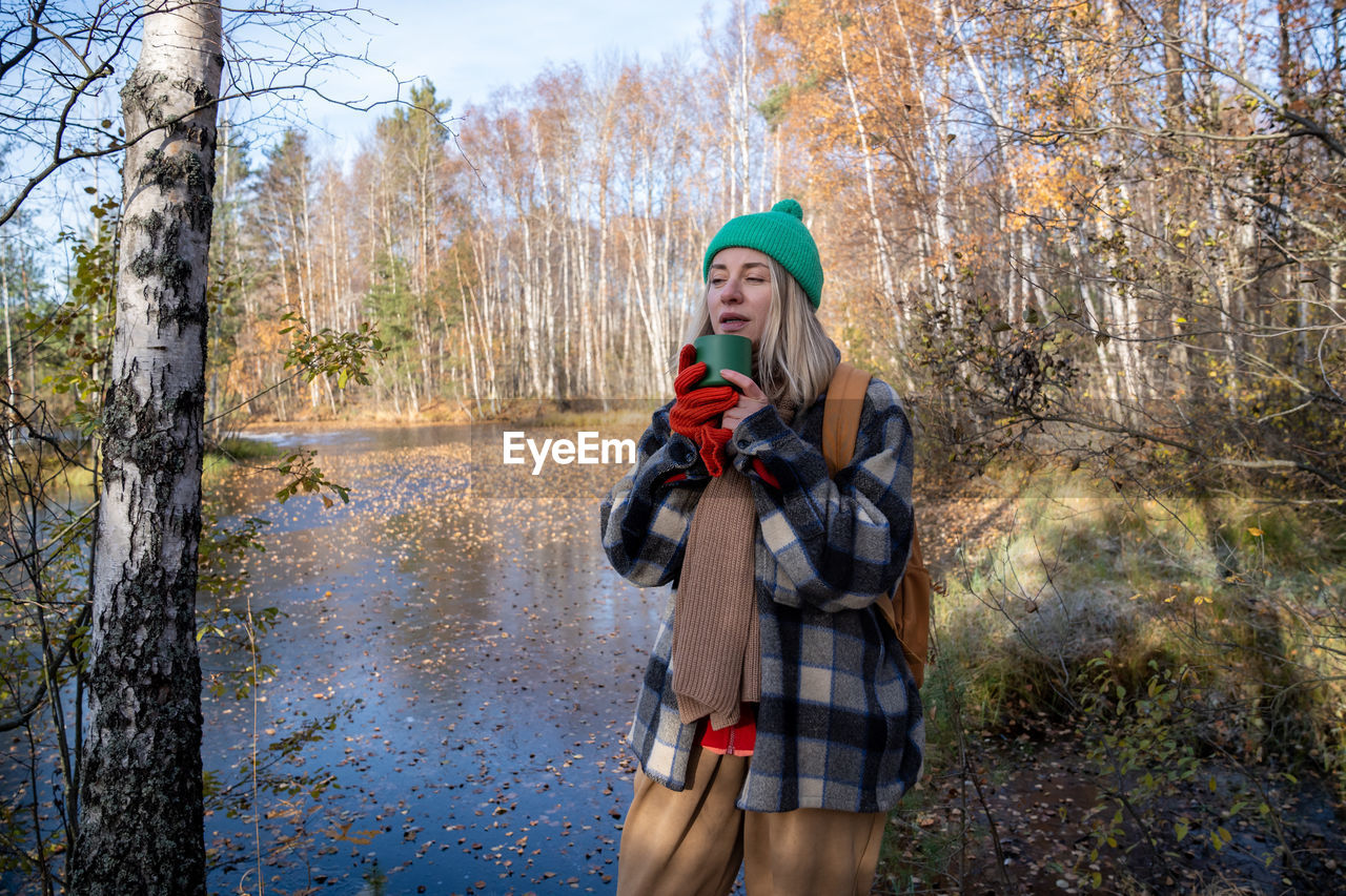 Freezing middle aged tourist woman hiker drinking hot tea from cup enjoying nature forest lake.