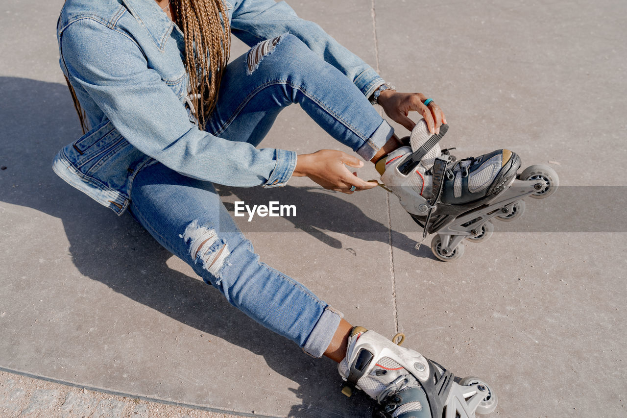 From above of african american female in denim outfit putting on rollerblades on sunny day in skate park