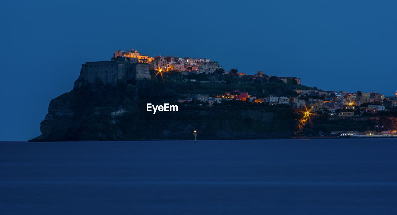 Illuminated building by sea against clear blue sky at night