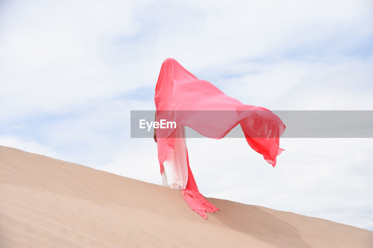 Caucasian woman standing in desert against blue sky, covered with pink fabric. face not visible. 