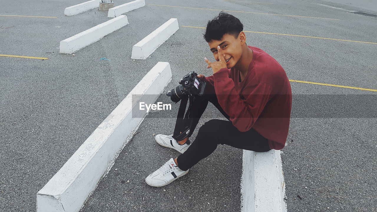 Portrait of smiling man with camera gesturing while sitting on barrier