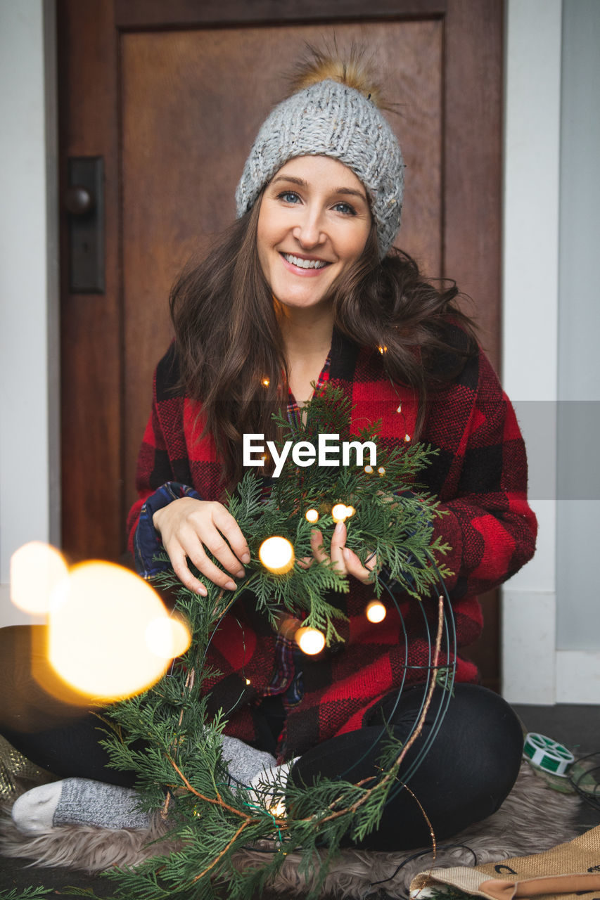 Portrait of smiling woman holding wreath while sitting at home during christmas