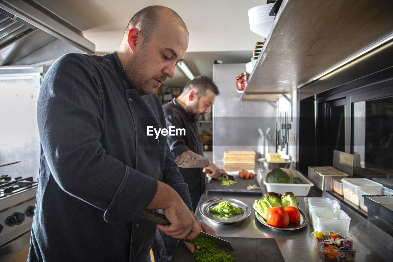 Side view of professional male cooks in uniforms cutting greens and vegetable while preparing food at counter in light kitchen of modern restaurant