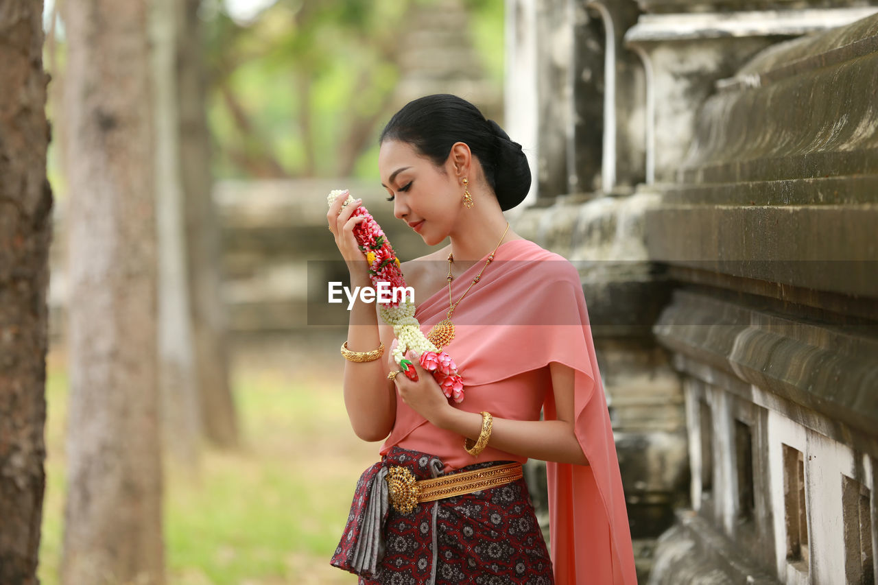 Young woman in traditional clothing holding flowers while standing against wall