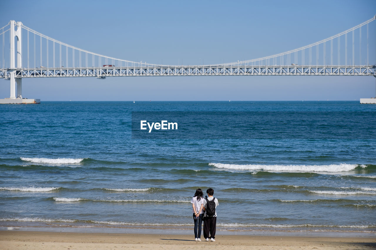 water, sea, bridge, suspension bridge, architecture, built structure, sky, beach, land, nature, travel destinations, horizon, travel, transportation, tourism, engineering, ocean, shore, beauty in nature, vacation, scenics - nature, two people, motion, horizon over water, coast, day, men, adult, wave, clear sky, holiday, outdoors, rear view, lifestyles, togetherness, women, city, trip, full length, pier, standing, body of water, copy space, walking, tranquil scene, tranquility, leisure activity