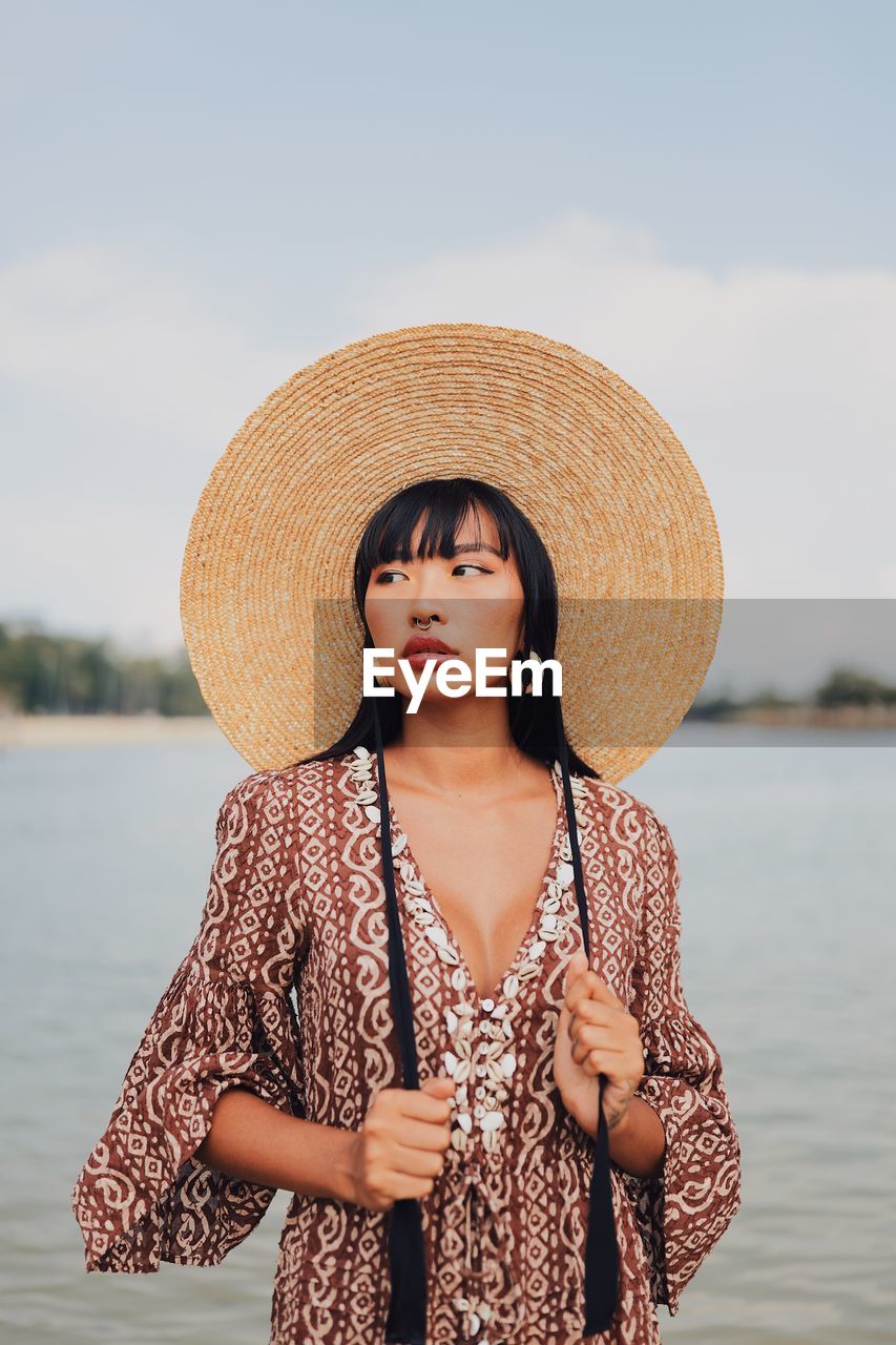 Young woman wearing hat looking away standing at beach against sky