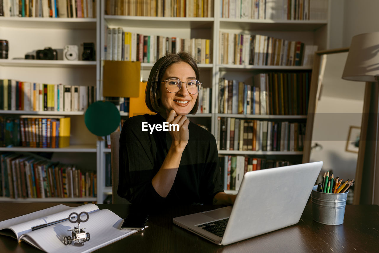 Businesswoman with hand on chin using laptop while sitting against bookshelf