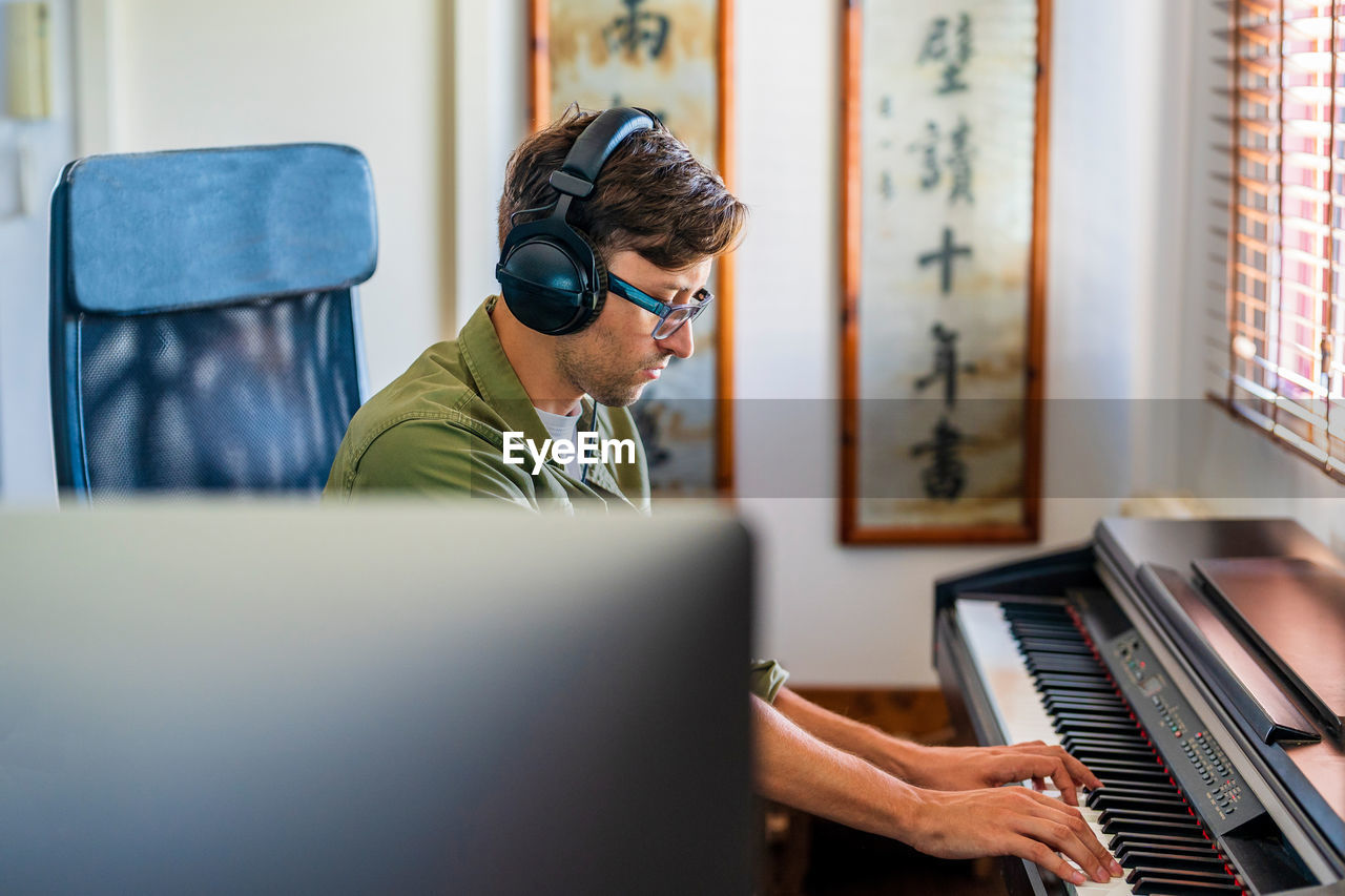 Side view of male musician in casual clothes playing melody on piano while sitting in modern apartment near window