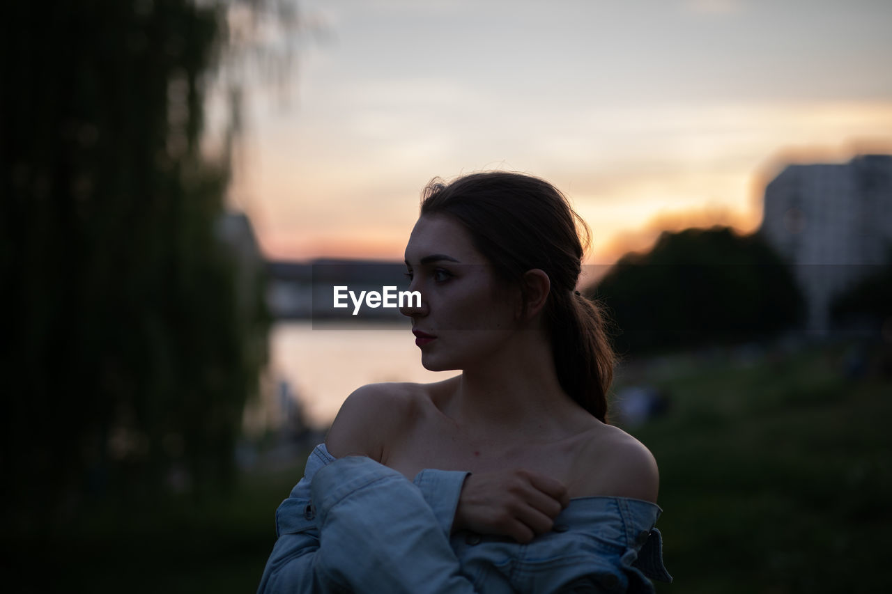 Young woman looking away while standing outdoors during sunset