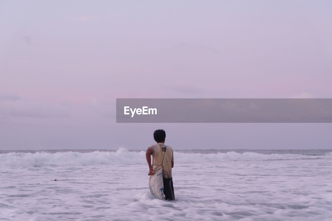 Rear view of man standing in sea against sky during sunset