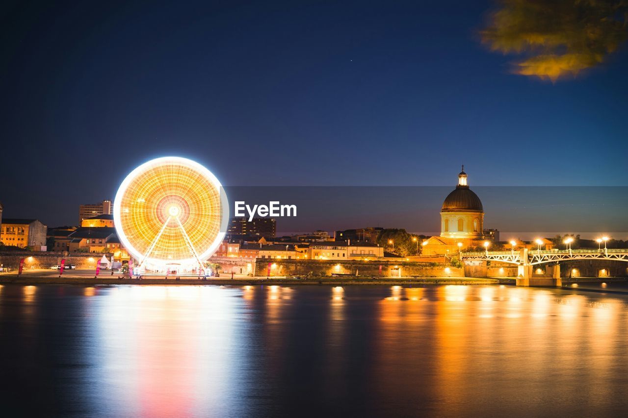 Illuminated ferris wheel and hospital de la grave by river against sky at dusk