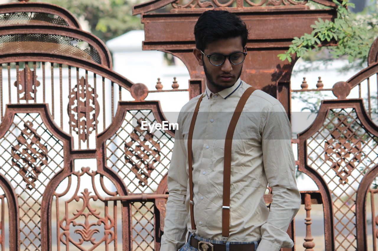 Thoughtful young man wearing suspenders standing against gate