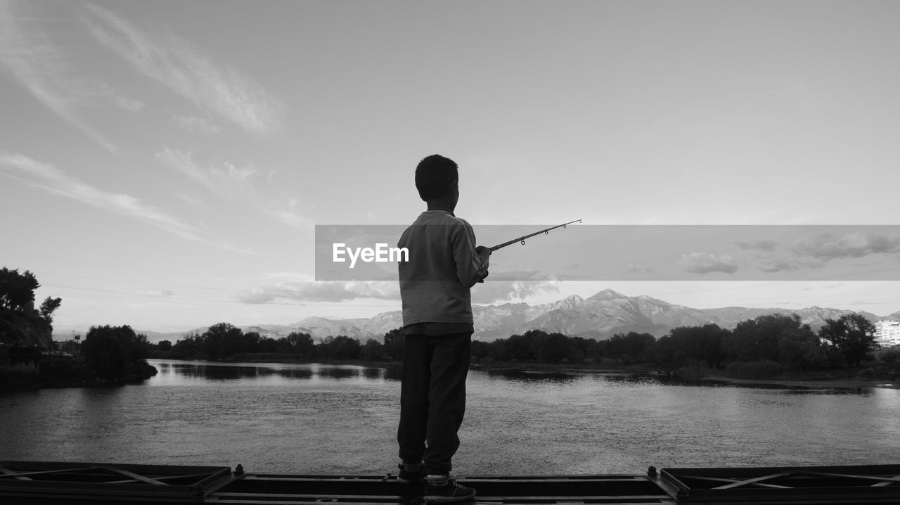 Rear view of boy fishing while standing on lake against sky