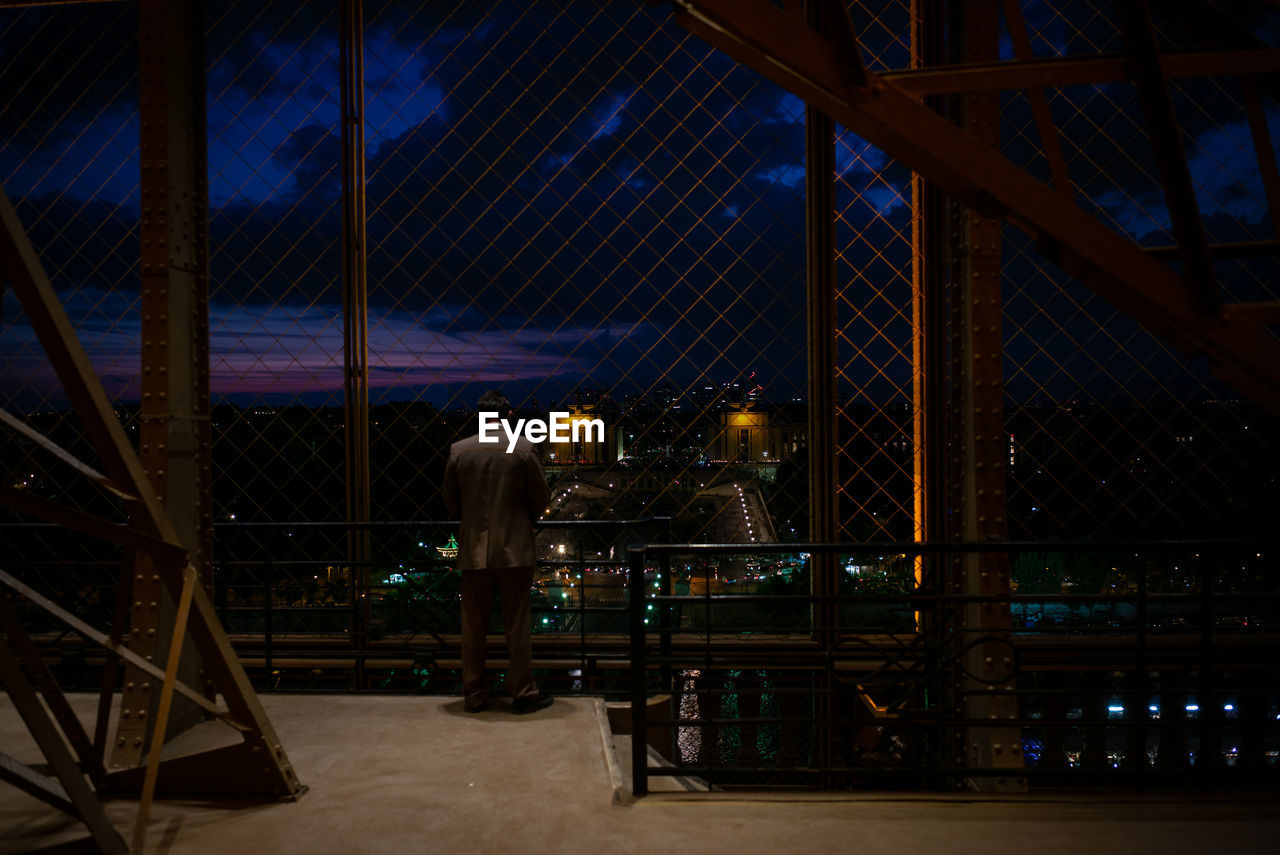 Rear view of man looking at city through fence at night