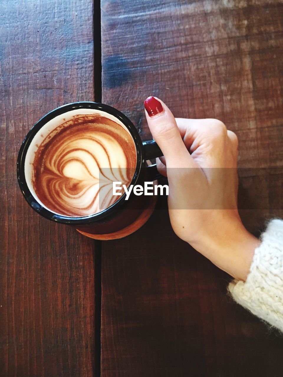 Cropped hand of woman holding cappuccino in cup on wooden table