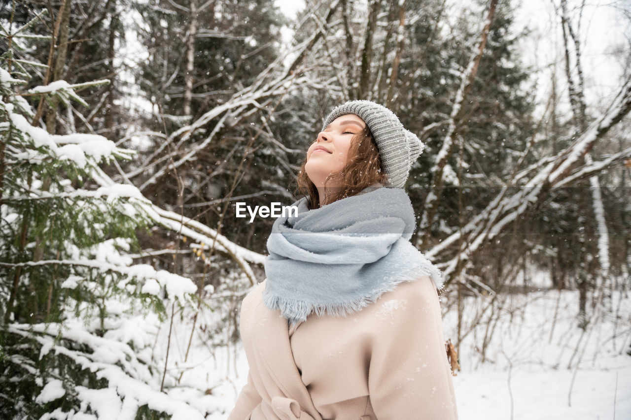 A cute girl stands under the snow with her eyes closed and smiles