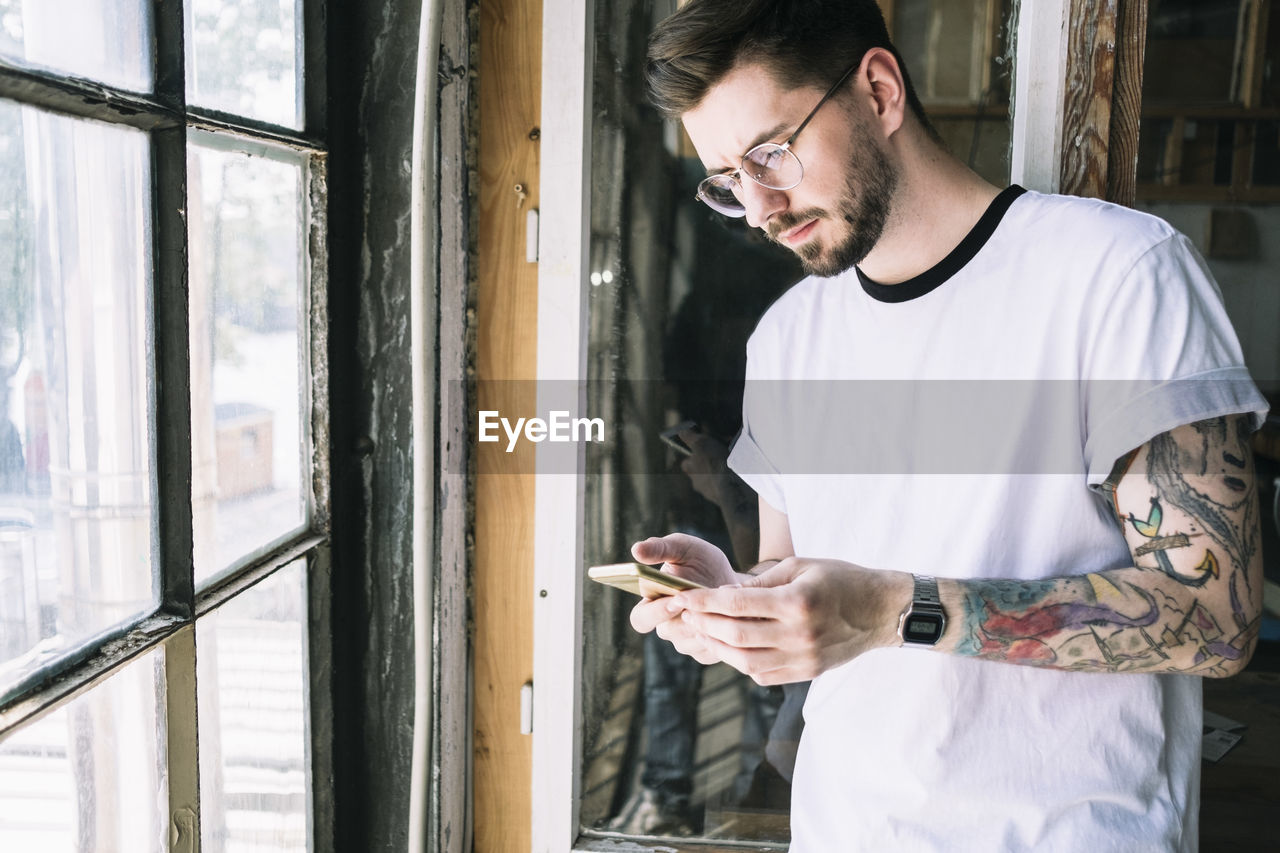 Confident male hipster hacker text messaging through smart phone by window at creative office