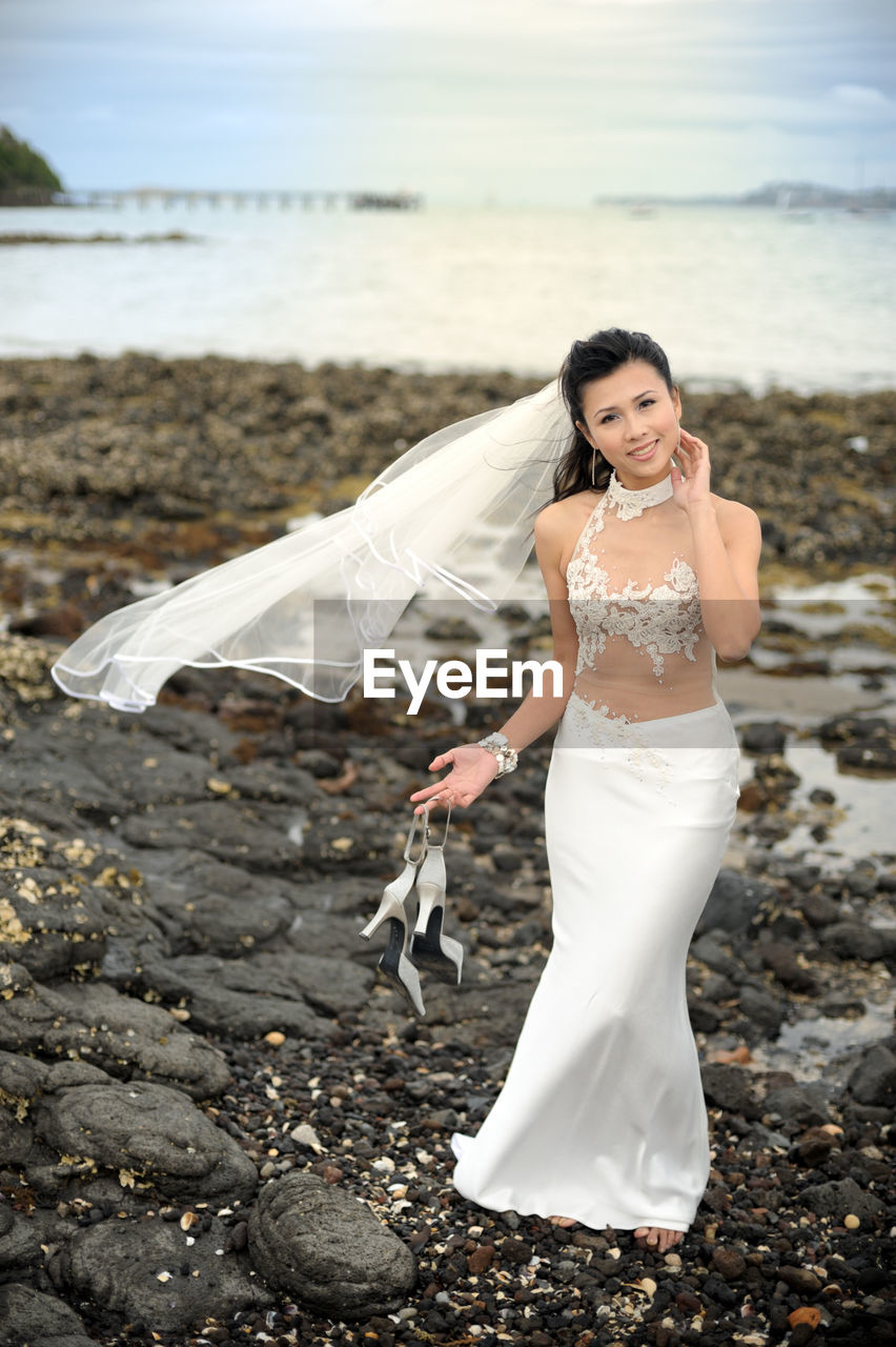 Full length portrait of smiling bride holding high heels while standing at beach