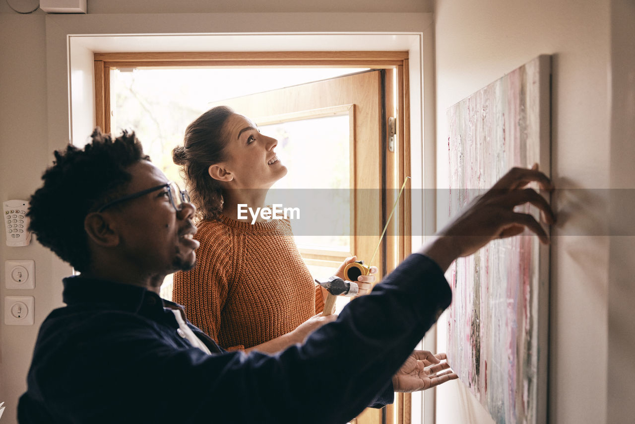 Couple assisting each other while hanging painting on wall near doorway at home